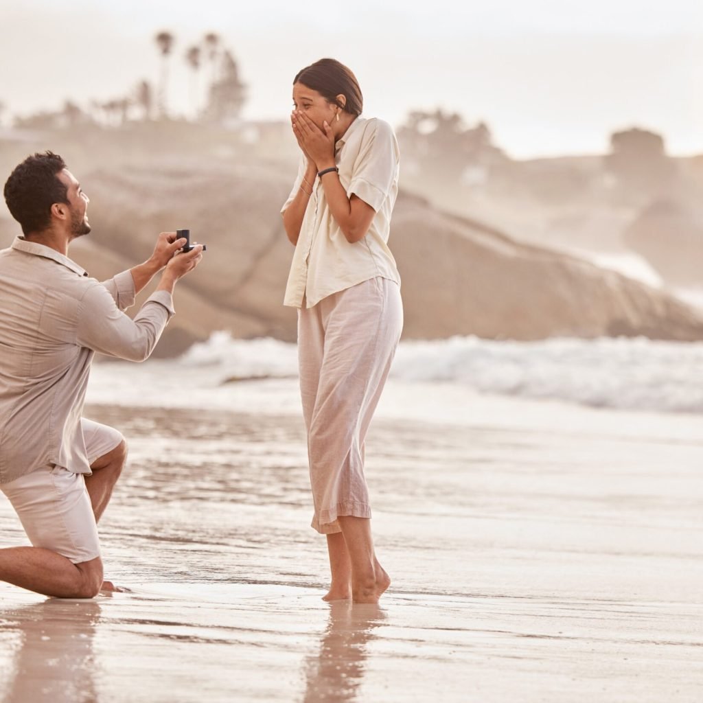 Couple at beach, surprise proposal and engagement with love and commitment with ocean and people outdoor. Travel, seaside and man propose marriage to woman, wow reaction and happiness with care.